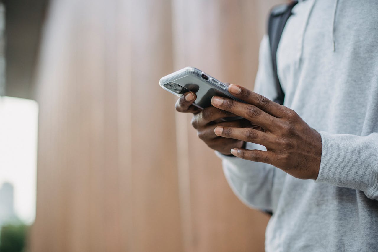 From below crop of unrecognizable African American young guy in casual clothes messaging on mobile phone while standing near wooden wall